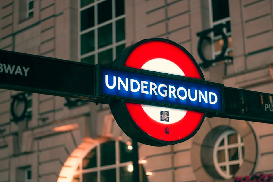 Close-up of a London Underground station sign displaying the word 'UNDERGROUND' in white letters on a blue background, mounted on a black pole with a red and white roundel logo. The sign is situated outside a building with stone walls and large windows, with a streetlamp casting light nearby. In the context of house removals or furniture transport, this signage may indicate the proximity of transportation links essential for efficient home relocation and moving logistics managed by Bow Movers. The image captures a part of an urban scene at dusk or nighttime, emphasizing the importance of transportation infrastructure in residential moving services, with the sign positioned centrally against the urban background.