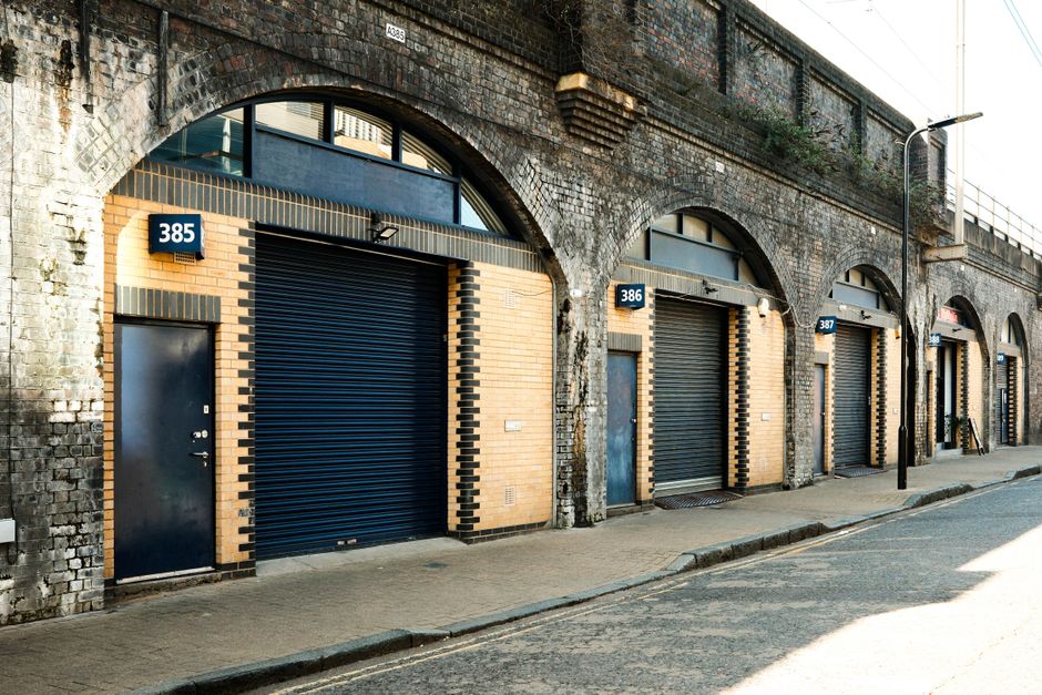 A row of four industrial-style commercial units with black roller shutter doors, each numbered 385, 386, 387, and 388, set into a brick and stone building with arched openings above. The units are situated along a quiet urban street with a pavement running parallel to their facades. Building materials include yellow-orange bricks, dark bricks creating decorative patterns around the doors, and weathered stonework above the arches. The second unit, labeled 386, is partially open revealing the entrance to a storage or industrial space, while the other doors are closed. To the right, a black bollard and a streetlamp are visible, with the street partly lit by sunlight. The environment appears clean and well-maintained, consistent with commercial property areas prepared for moving or loading activities, which Bow Movers could facilitate as part of home relocation or furniture transport services.
