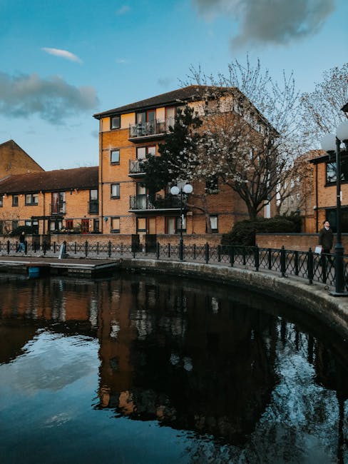 A riverside scene featuring modern and traditional residential buildings with distinctive architectural details, situated along a calm waterway. The buildings are illuminated by exterior lighting, wit