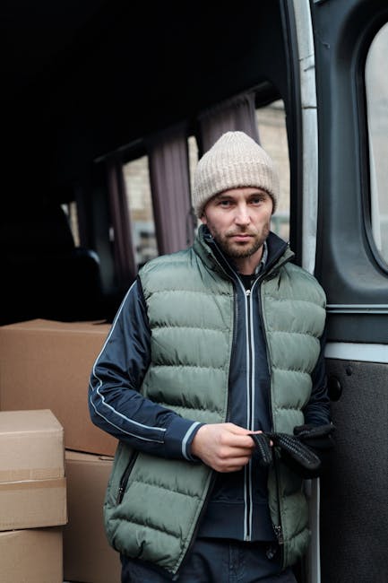 A man dressed in a grey knit beanie hat, navy and green puffer jacket, and dark clothing stands inside a moving van, preparing for a home relocation. Behind him, several packed cardboard boxes are stacked, likely filled with household items for packing and moving. The van’s interior is visible with dark fabric curtains and a window, indicating a professional removal or moving service. The man appears focused as he handles a strap or cord, possibly used for securing furniture or boxes during transport. This scene captures an active loading process, with the environment well-lit and organized for furniture transport and a smooth moving operation. The presence of packing materials and the vehicle's setup reflect professional house removals, exemplifying logistics involved in furniture transport and home relocation, as services provided by Bow Movers.
