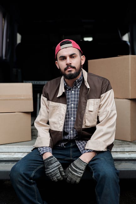 A man dressed in a grey knit beanie hat, navy and green puffer jacket, and dark clothing stands inside a moving van, preparing for a home relocation. Behind him, several packed cardboard boxes are stacked, likely filled with household items for packing and moving. The van’s interior is visible with dark fabric curtains and a window, indicating a professional removal or moving service. The man appears focused as he handles a strap or cord, possibly used for securing furniture or boxes during transport. This scene captures an active loading process, with the environment well-lit and organized for furniture transport and a smooth moving operation. The presence of packing materials and the vehicle's setup reflect professional house removals, exemplifying logistics involved in furniture transport and home relocation, as services provided by Bow Movers.