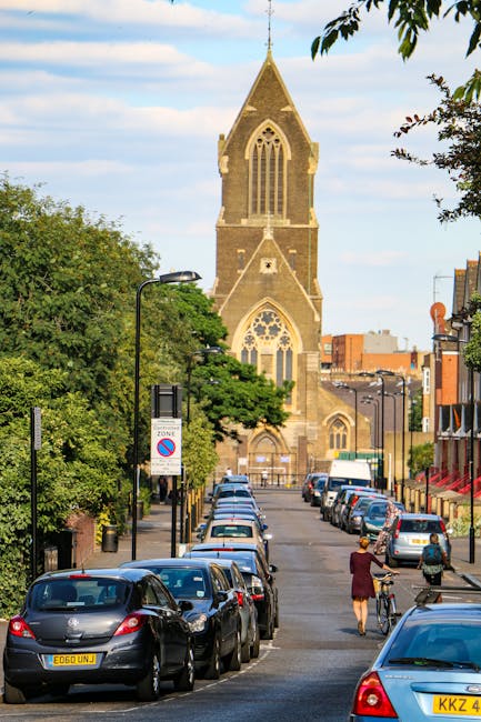 A street scene showing a busy residential area with parked cars along the pavement and trees lining the roadside. In the background, a historic clock tower with a pointed roof and ornate architectural