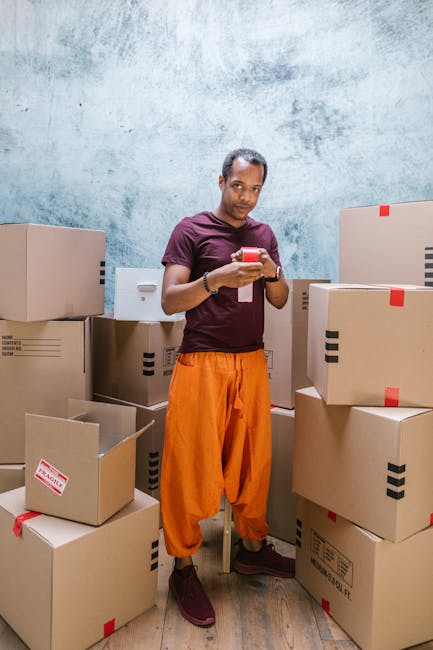 A man dressed in an orange pair of trousers, a maroon T-shirt, and brown shoes is standing indoors, surrounded by multiple cardboard boxes of various sizes, some sealed with red tape and others open,