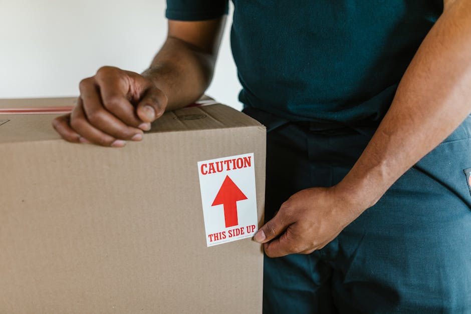 A man dressed in an orange pair of trousers, a maroon T-shirt, and brown shoes is standing indoors, surrounded by multiple cardboard boxes of various sizes, some sealed with red tape and others open,